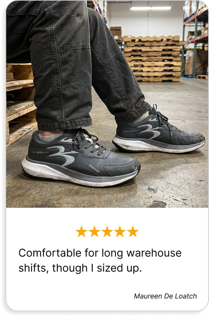 Dark grey Gdefy athletic shoes with white soles, worn by a person standing in a warehouse.