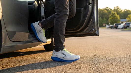 A driver steps out of a car onto rest-stop asphalt wearing grey G-Defy Mighty Walk walking shoes with blue VersoShock soles