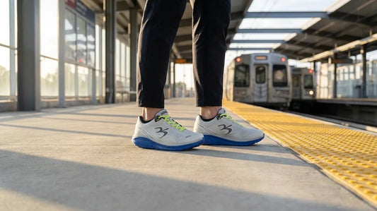 Ground-level view of a commuter's shoes for commuting standing on a train platform beside the yellow safety strip, with a train arriving in the background