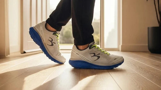 Close view at floor level of an adult mid-stride in grey and blue G-Defy Mighty Walk walking shoes on a sunlit wooden floor