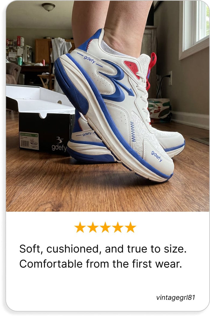 Gdefy athletic shoe in white, blue, and red, worn on a person's foot, with the shoe box nearby on a wooden floor.