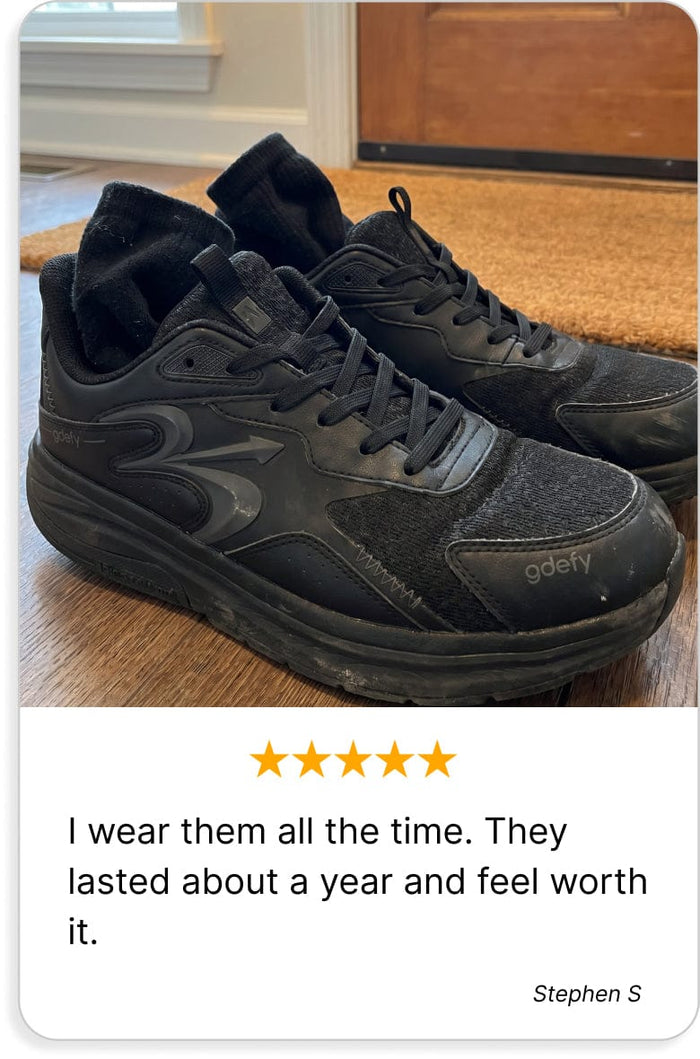Black Gdefy sneakers with black socks inside, appearing worn, on a wooden floor near a doormat.
