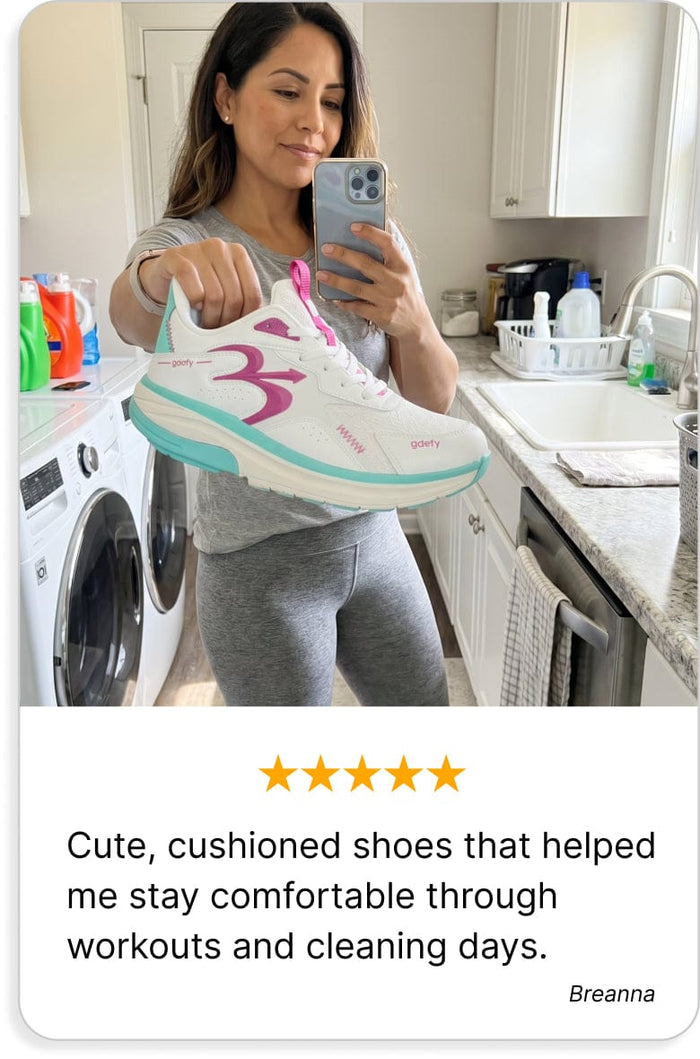 White, pink, and teal GDefy sneaker held by a woman taking a mirror selfie in a laundry room.