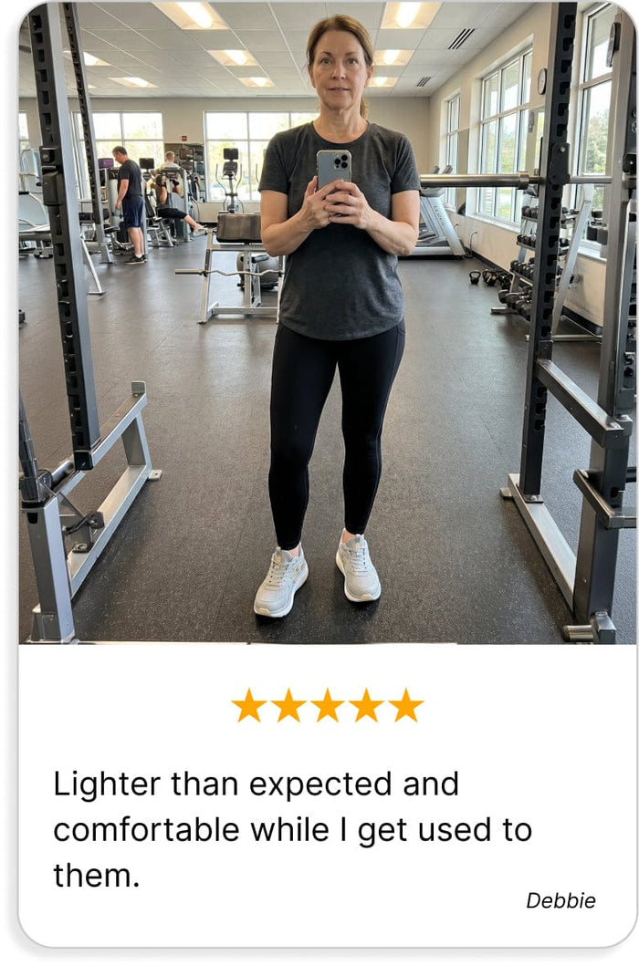 Athletic shoes, light grey with white soles and subtle pink accents, worn by a woman taking a mirror selfie in a gym.