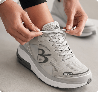 Light gray GDEFY running shoe with white sole and dark gray accents, being tied by a person.