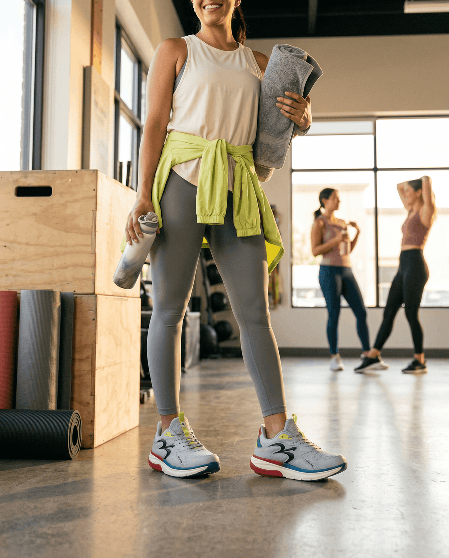 GDEFY Energiya crossfit shoes in gray, lime, red, and blue worn by a woman holding a water bottle and towel in a gym.